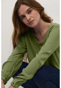 Ung kvinna med långt brunt hår, klädd i en strukturerad grön långärmad tröja och blå jeans, sittande mot en enkel ljus bakgrund.