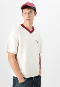 Pulover tricotat cu umeri scurți de culoare crem, cu un guler în V ribat de culoare vișinie și logo brodat. Purtați-l cu o șapcă roșie decolorată, asortată cu blugi închisi.