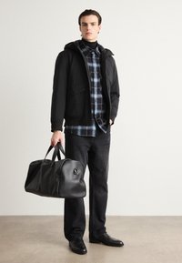 Μαύρο μπουφάν με κουκούλα πάνω από ένα μπλε και μαύρο καρό πουκάμισο, συνδυασμένο με σκούρα τζιν και μαύρα δερμάτινα παπούτσια, κρατώντας μια μαύρη τσάντα γυμναστηρίου.