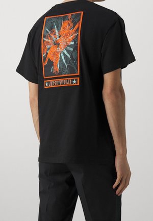 Homme portant un t-shirt noir avec un tigre orange, une figure ailée et le texte "JUST WILD" dans le dos.