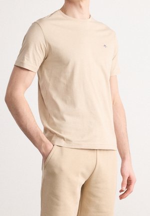 Mężczyzna ubrany w gładki beżowy T-shirt z krótkim rękawem i dopasowane beżowe spodnie, z jedną ręką w kieszeni, na jednolitym tle.