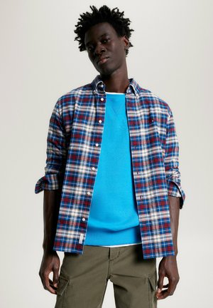 Camicia a scacchi blu, rossa e bianca; sovrapposta a un maglione blu uniforme. L'outfit è completato da pantaloni cargo verde oliva.