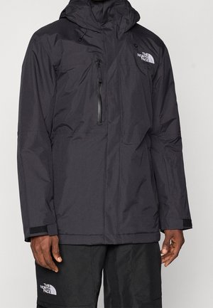 Mężczyzna ubrany w czarną wodoodporną kurtkę North Face z kapturem i kieszenią na piersi na zamek błyskawiczny, w połączeniu z czarnymi spodniami North Face.