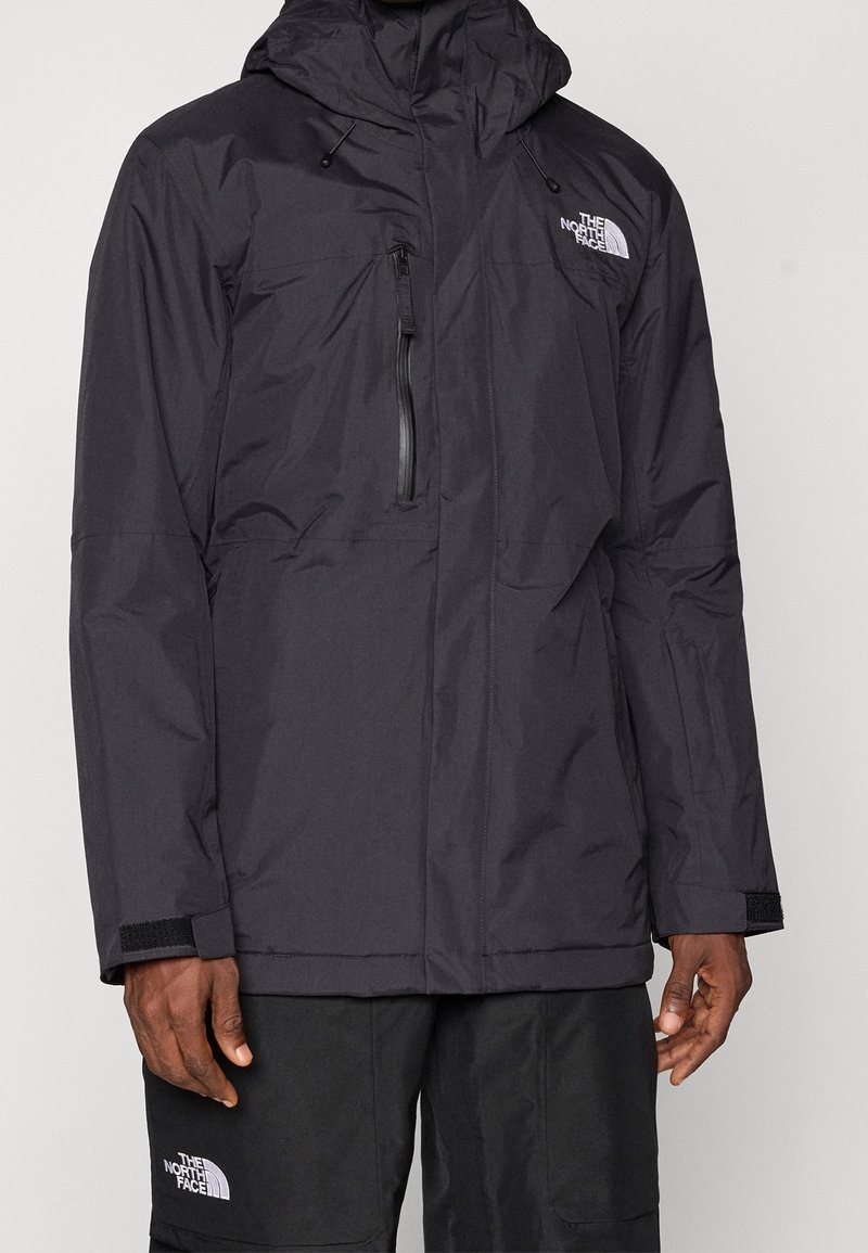 Homme portant une veste imperméable noire North Face avec capuche et poche poitrine zippée, associée à un pantalon noir North Face.