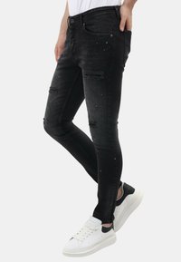 Smalle zwarte jeans met een distressed uitstraling, voorzien van kleurrijke verfspetters. Gecombineerd met witte sneakers met zwarte accenten.