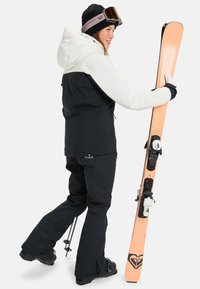 Narciarz ma na sobie czarną i białą wodoodporną kurtkę, czarne spodnie narciarskie i trzyma pomarańczową nartę z czarnymi akcentami. Ma na sobie czarne rękawice i gogle.