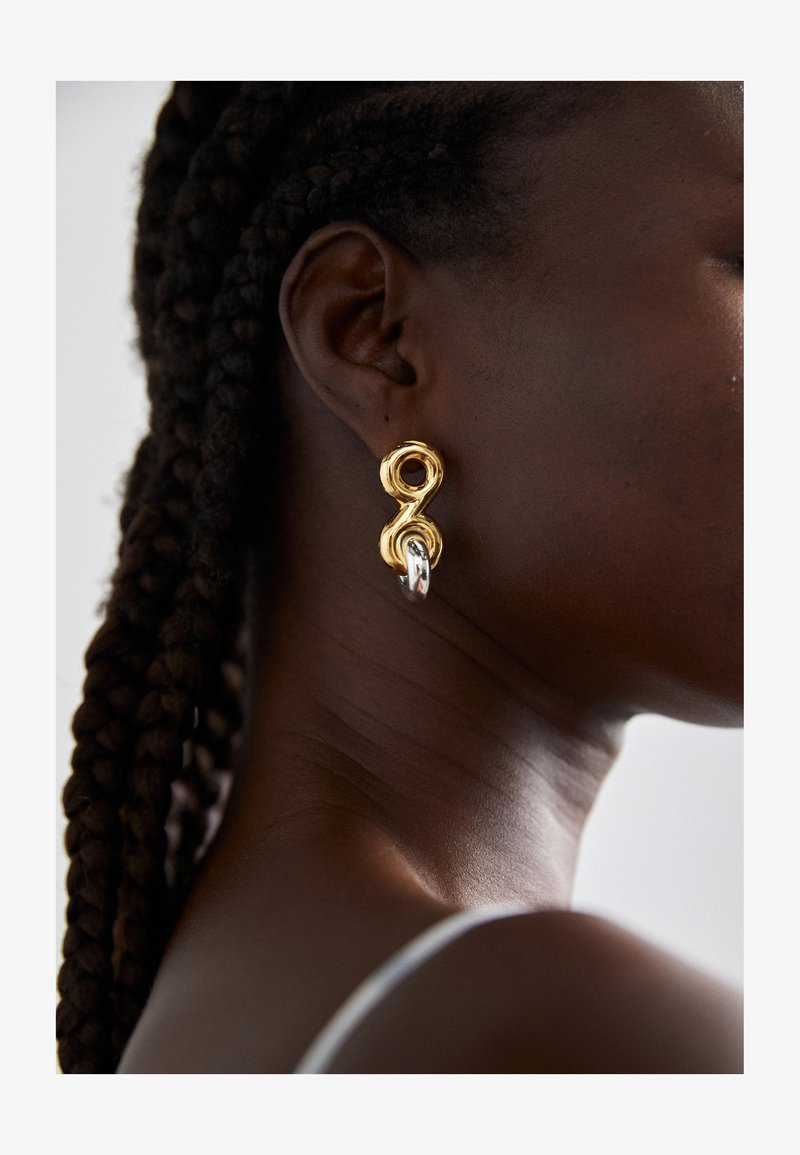 Boucles d'oreilles abstraites en or et argent avec un design lisse et torsadé. Elles présentent une boucle proéminente et une finition texturée.