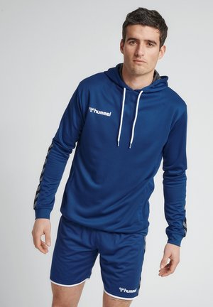 Haut de sport bleu marine avec des rayures grises sur les manches, logo sur la poitrine, associé à un short assorti avec une bordure blanche. Texture de tissu lisse.