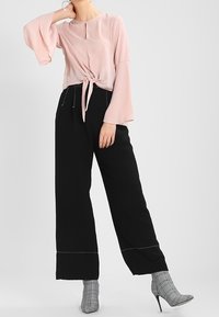 Vaaleanpunainen nauhoitettu paita, jossa on pitkät kellohihat, yhdistettynä korkeavyötäröisiin mustiin leveälahkeisiin housuihin, joissa on vaaleat tikkaukset, sekä ruutukuvioisiin korkokenkiin.