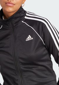 Black zip-up sports jacket with white stripes on shoulders, featuring an embroidered Adidas logo on the chest. Smooth, lightweight fabric.