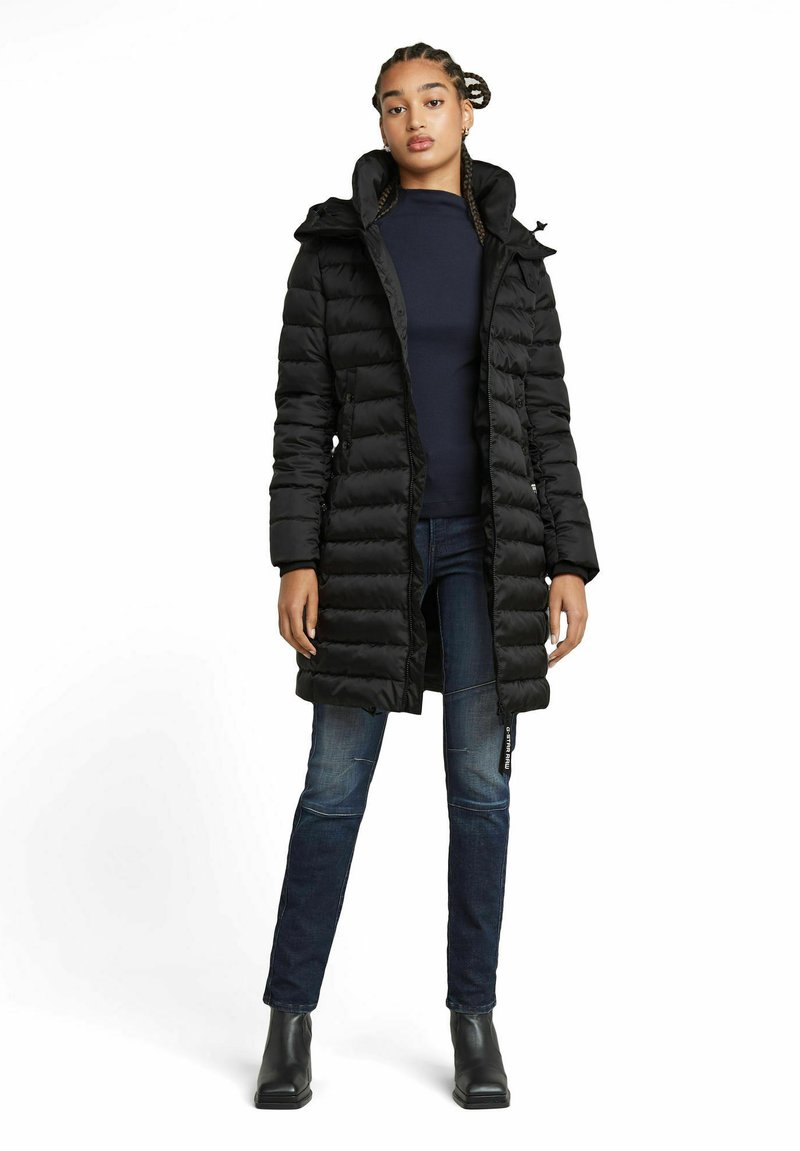 Schwarze Daunenjacke mit Kapuze, mit gestepptem Design, Frontreißverschluss und Seitentaschen. Tragen über einem dunklen Oberteil und blauen Jeans.