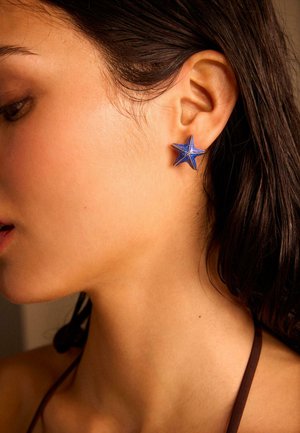 Žensko lijevo uho s plavom naušnicom u obliku zvijezde, tamna kosa jeza ušima, a na ramenu se vidi smeđi remen.