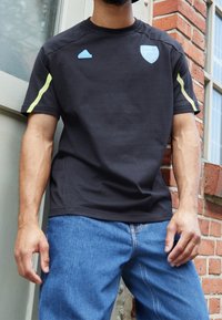 Černé tričko s krátkým rukávem s světle modrým logem Adidas a znakem Arsenalu. Neonově žluté akcenty na ramenou. Volný střih s hladkou strukturou.