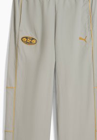 Jasnoszare spodnie dresowe PUMA o błyszczącej teksturze, z owalnym logo i żółtymi akcentami wzdłuż szwów.