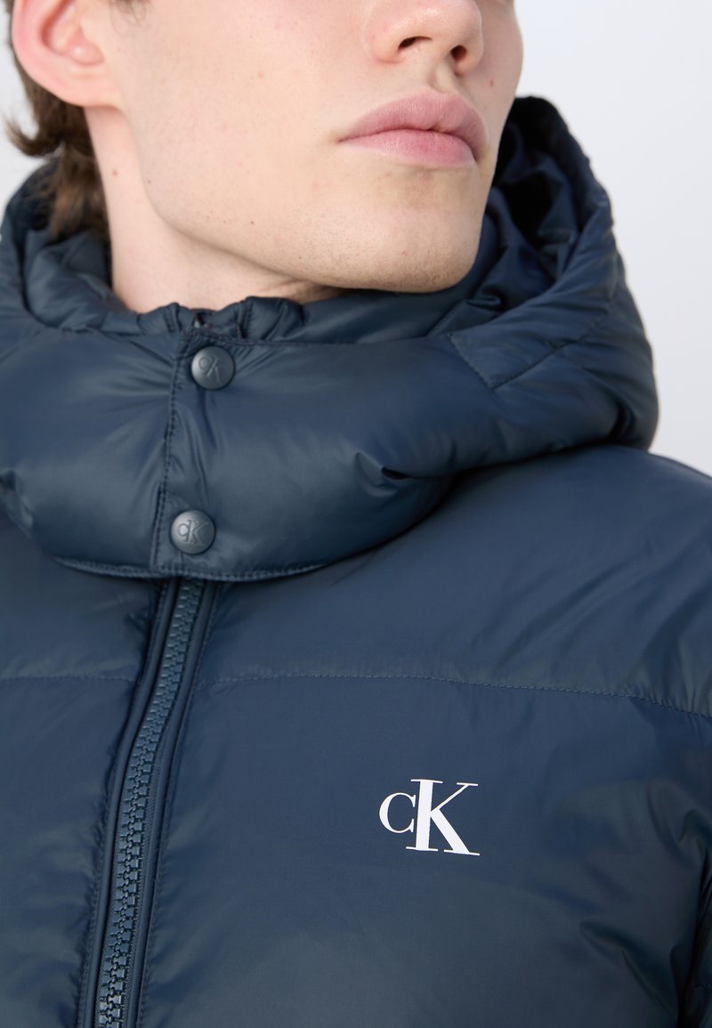 Puffermantel in Dunkelblau, mit hohem Kragen, sichtbarem Reißverschluss und Knopfdetails. Logo in Weiß auf der linken Brustseite.