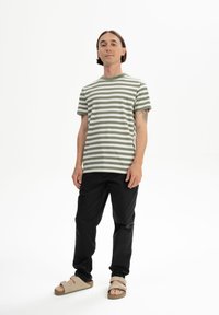 Gestreiftes T-Shirt in hellem Olivgrün und Weiß, kombiniert mit schwarzen Hosen und beigen Sandalen. Einfaches Design, weicher Stoff und lockere Passform.