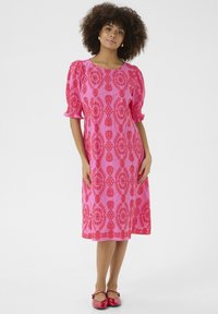 Roza obleka s puhastimi rokavi in geometrijskim rdečim vzorcem. A-linijska silhueta s valovitim robom in gladko teksturo. Kombinirana z rdečimi balerinkami.