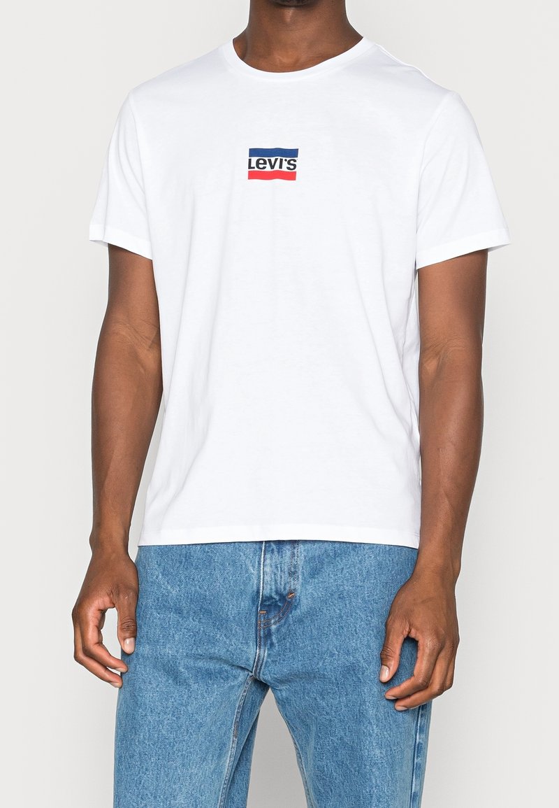 Homme portant un t-shirt blanc Levi's avec un logo bleu et rouge, associé à un jean taille haute bleu clair.
