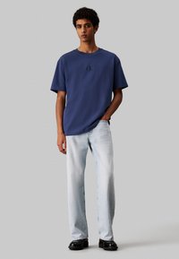 Camiseta de algodón azul marino con mangas cortas, con un logo bordado. Combinada con jeans rectos de color azul claro y zapatos negros.