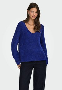 Modra, teksturirana pulover s globokim V-izrezom in dolgimi rokavi, izdelan iz mehkega, puhastega materiala, v kombinaciji s temnimi kavbojkami.