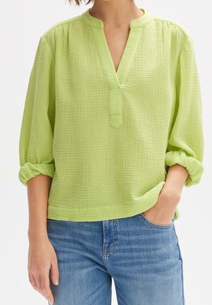 Femme portant un chemisier vert clair texturé à manches longues retroussées et un jean bleu, debout avec une main dans la poche.