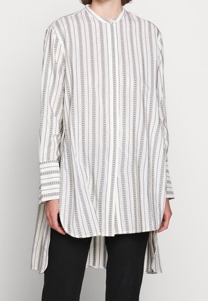 Woman wearing a long white button-up shirt with thin black vertical dotted stripes and black pants against a plain background.