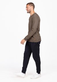 Camicia a maniche lunghe marrone in tessuto strutturato, abbinata a pantaloni jogger neri e sneakers bianche, in piedi di fronte a uno sfondo bianco uniforme.