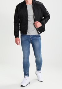 Giubbotto bomber nero con zip frontale, t-shirt grigia, jeans slim blu e sneakers bianche. Il giubbotto ha tasche laterali e polsini a coste.
