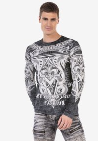 Schwarzes Langarmshirt mit aufwendigen weißen grafischen Designs von Schlangen und einer Fleur-de-Lis, kombiniert mit abgenutzten grauen Jeans.