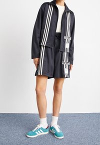 Svart idrottsjacka och shorts med vita ränder, utrustad med en logotyp-patch. Teal skor med vita sulor och detaljer, bärs med vita strumpor.