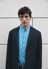 Sort blazer båret over en lyseblå skjorte med knapper. Modellen står foran en tekstureret, lys væg. Nærbillede med fokus på tøjet.