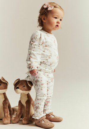 Peuter in bloemenpyjama en een roze strik staand naast twee bruine knuffelhondjes op een neutrale achtergrond.