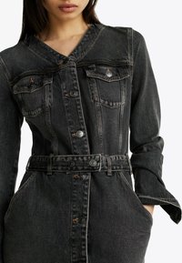 Abito in denim scuro con chiusura a bottoni, due tasche sul petto e vita con cintura. Presenta un scollo a V e maniche lunghe. Tessuto testurizzato.
