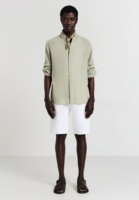 Camicia button-up di colore verde chiaro con pieghe, pantaloni corti bianchi e sandali marroni. Una sciarpa beige aggiunge un tocco di stile. Il modello è in piedi davanti a uno sfondo neutro.