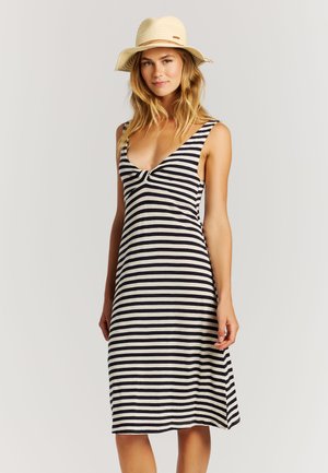 Vrouw die een zwart-wit gestreepte mouwloze jurk en een beige zonnenhoed draagt, staand tegen een effen lichte achtergrond.