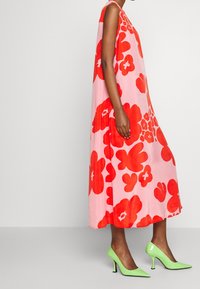 Vestido midi sin mangas en rosa con grandes patrones florales rojos. Combinado con zapatos de tacón altos brillantes en verde. Tela suave, corte relajado.