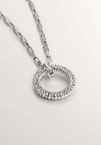 Collar de cadena de plata con un colgante circular incrustado con piedras preciosas transparentes y brillantes sobre un fondo blanco.