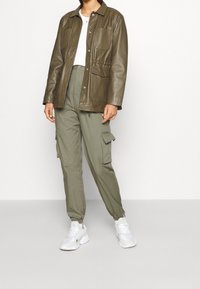 Brun läderjacka med framfickor, i kombination med olivgröna cargobyxor och vita sneakers. Slät textur, avslappnad passform och knappstängningar.