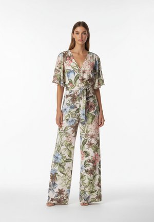 Frau trägt einen weißen, floralen Jumpsuit mit weitem Bein, V-Ausschnitt, flatternden Ärmeln und gebundenem Taillengürtel, stehend vor einem einfarbigen Hintergrund.