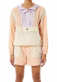 Kleurgeblokte windbreaker in zachte pastel tinten van roze, crème en lavendel, met een verstelbare capuchon en een voorkant zak. Gecombineerd met bijpassende shorts.