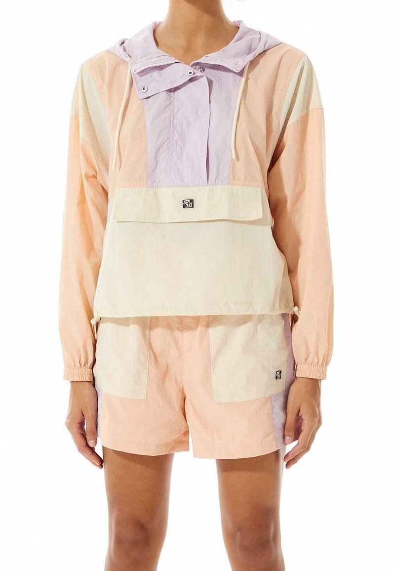 Kleurgeblokte windbreaker in zachte pastel tinten van roze, crème en lavendel, met een verstelbare capuchon en een voorkant zak. Gecombineerd met bijpassende shorts.