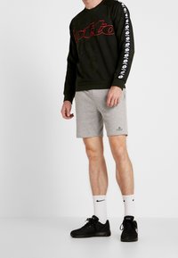 Uomo che indossa una felpa nera Lotto con logo e pantaloni corti grigi della marca First, in piedi su uno sfondo bianco.