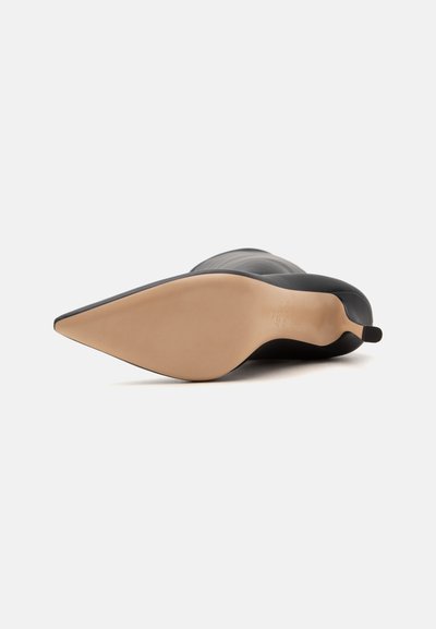 Bottine en cuir noir à bout pointu avec une semelle beige, vue de dessous à l'avant sur fond blanc.