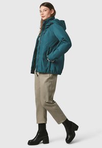 Giacca con cappuccio color teal, chiusura con zip, orlo elastico, due tasche laterali, abbinata a pantaloni beige e stivaletti neri con suola piatta.