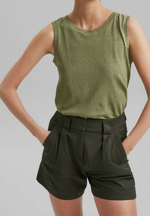 Femme portant un haut sans manches vert olive et un short taille haute vert foncé, mains dans les poches, sur un fond uni.