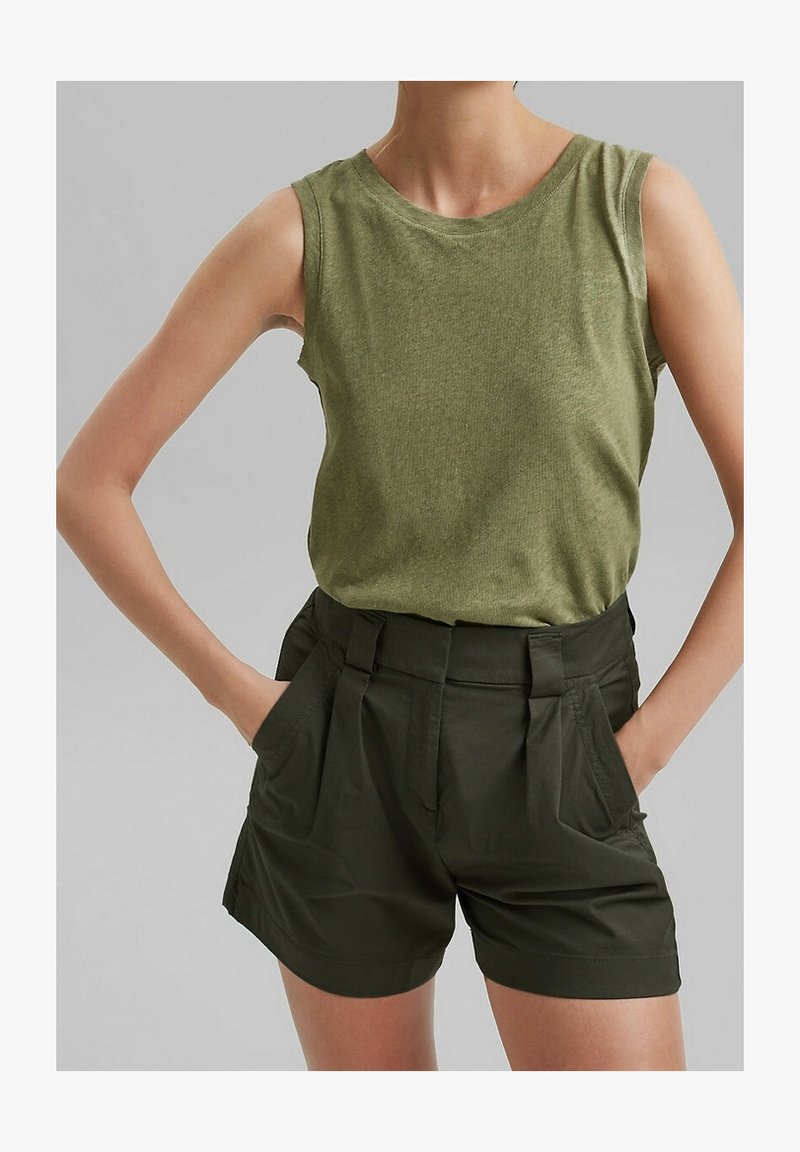 Femme portant un haut sans manches vert olive et un short taille haute vert foncé, mains dans les poches, sur un fond uni.