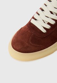 Brune semskede sneakers med rund tå, kremfargede snører og lys gummisåle. Har stikkede detaljer og glatt tekstur.