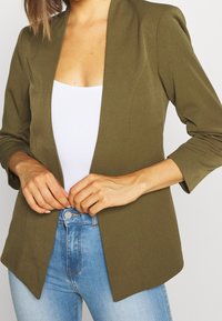 Olive green blazer with a structured design, sharp collar, and three-quarter sleeves, paired with a white tank top and light blue jeans.