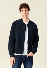 Chaqueta bomber de color navy con cremallera frontal, puños y cinturilla acanalados, llevada sobre una camisa blanca y combinada con jeans azules. Textura suave.