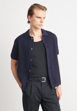 Navy ribbed short-sleeve cardigan with a collared neck, featuring brown buttons and a straight hem, worn over a black tank top and trousers.
